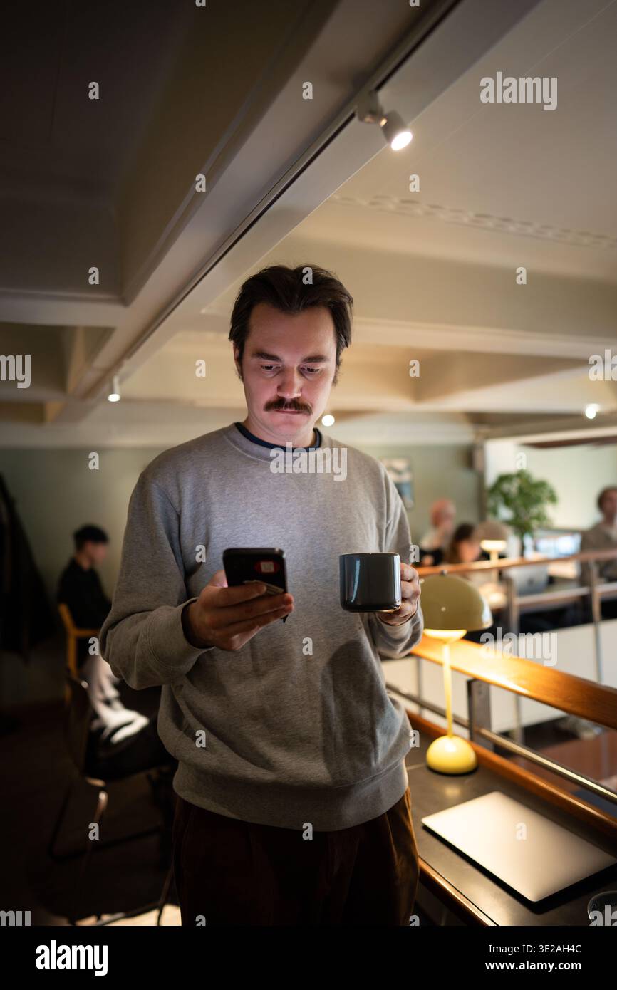 Handsome businessman enjoying coffee and scrolls smartphone after finishing work at co-working cafe. Stock Photo