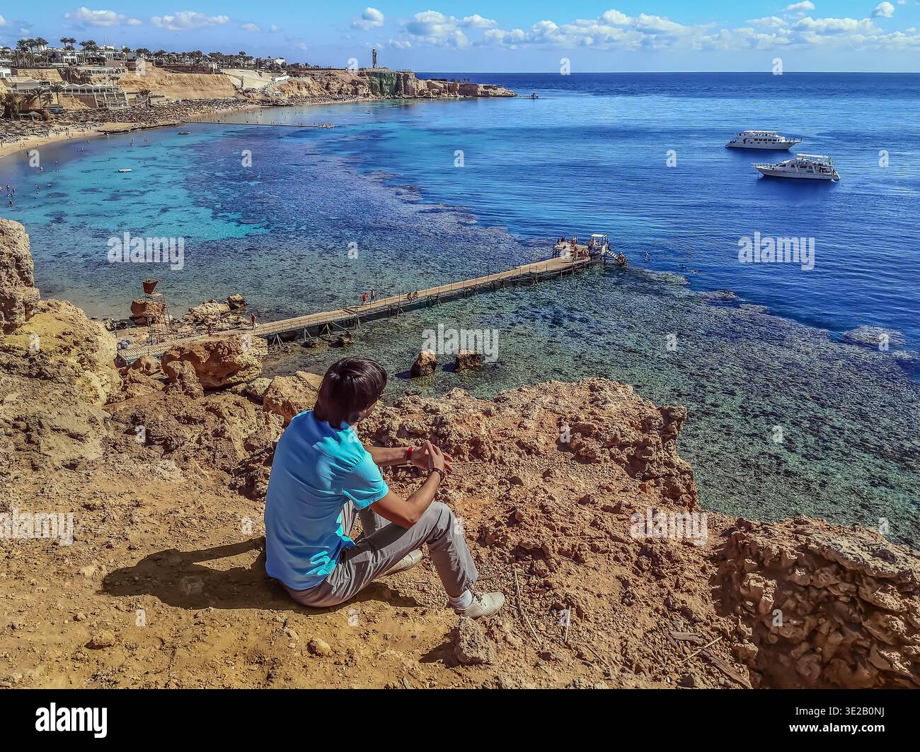 A man sitting on a rock looking down at the Red Sea with a coral reef in Sharm El Sheikh, Egypt - view from the back. An adult guy in front of a sea - Smartphone Captured Stock Image