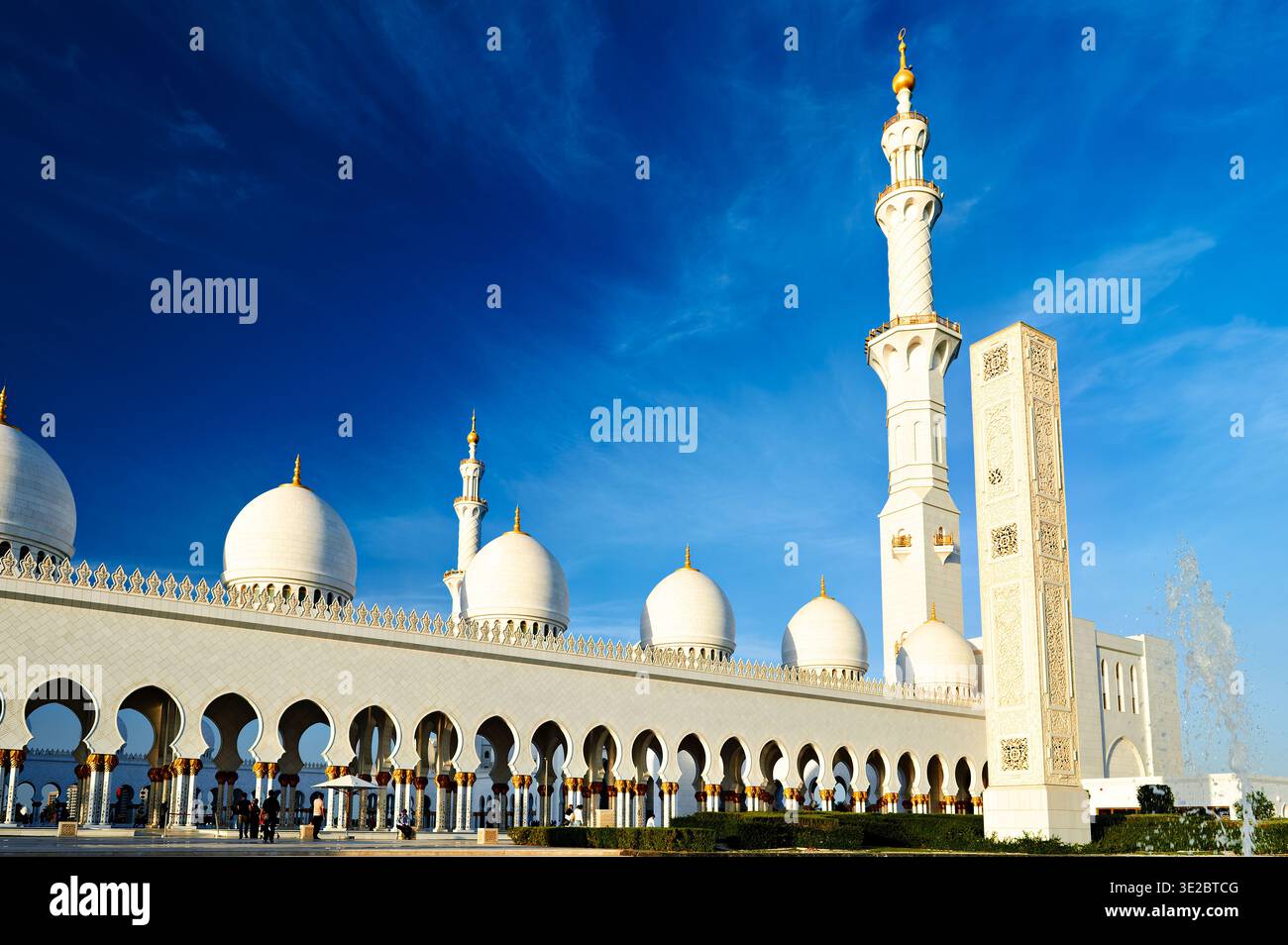 People explore the Sheikh Zayed Grand Mosque in Abu Dhabi under a clear blue sky. The mosque showcases its beautiful architecture. Stock Photo
