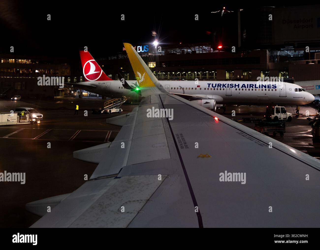 View from a passenger window over the wing to a Turkish Airlines aircraft on a wet night apron with ground service vehicles and lit terminal at Düssel Stock Photo