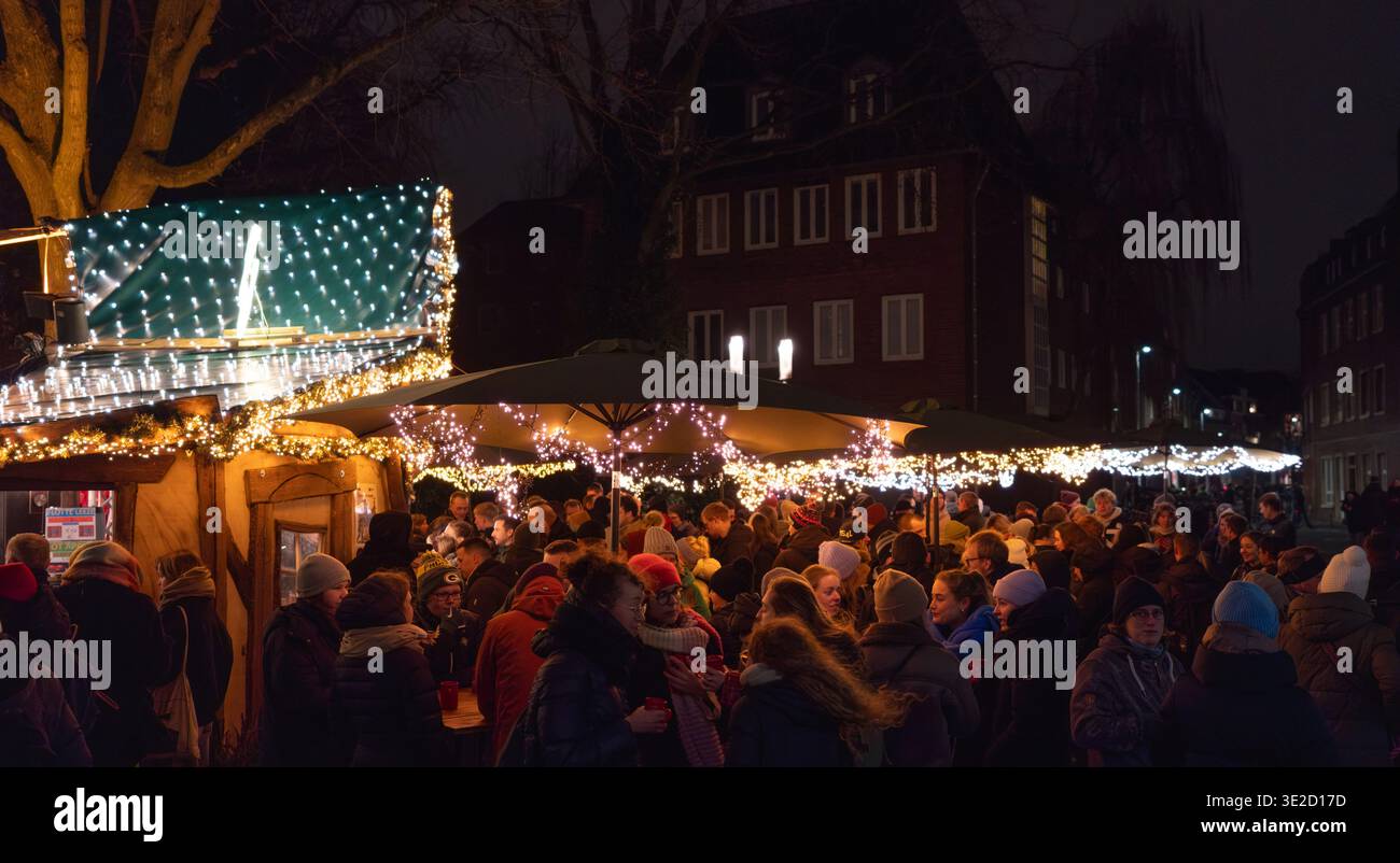 Crowded winter night market scene with warm fairy lights outdoor umbrellas and bundled visitors socializing at festive stalls. Munster, Germany Stock Photo