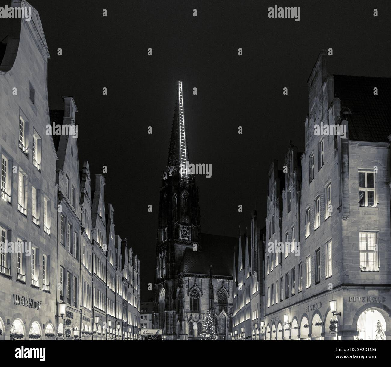 Moody black and white night scene of a narrow European old town street framed by historic buildings and a tall illuminated Gothic church spire with gl Stock Photo