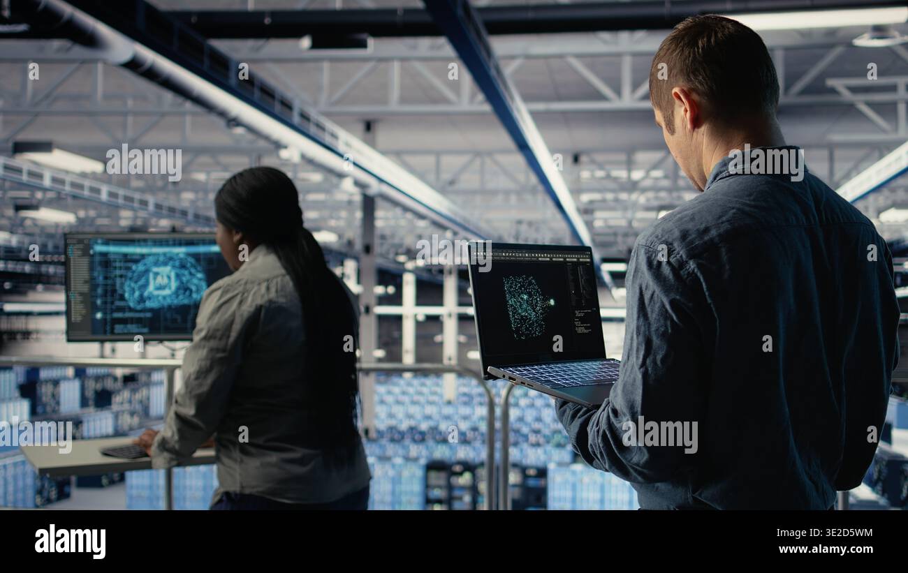 Software developer in data center checks AI dashboard using laptop during routine systems review. Server hub IT specialist tests artificial intelligence on notebook to check performance, camera A Stock Photo