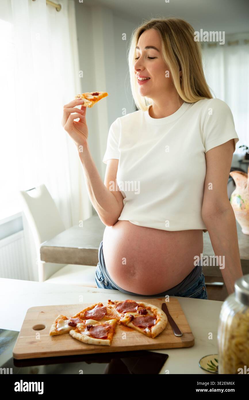 Happy pregnant woman eating pizza at home. Pregnancy cravings, maternity lifestyle, nutrition and prenatal wellness concept. Stock Photo