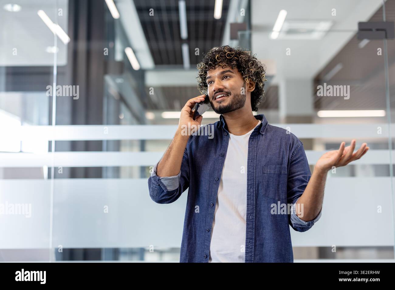 Smiling Indian man standing in the office and emotionally talking on the phone, gesturing with his hands. Stock Photo