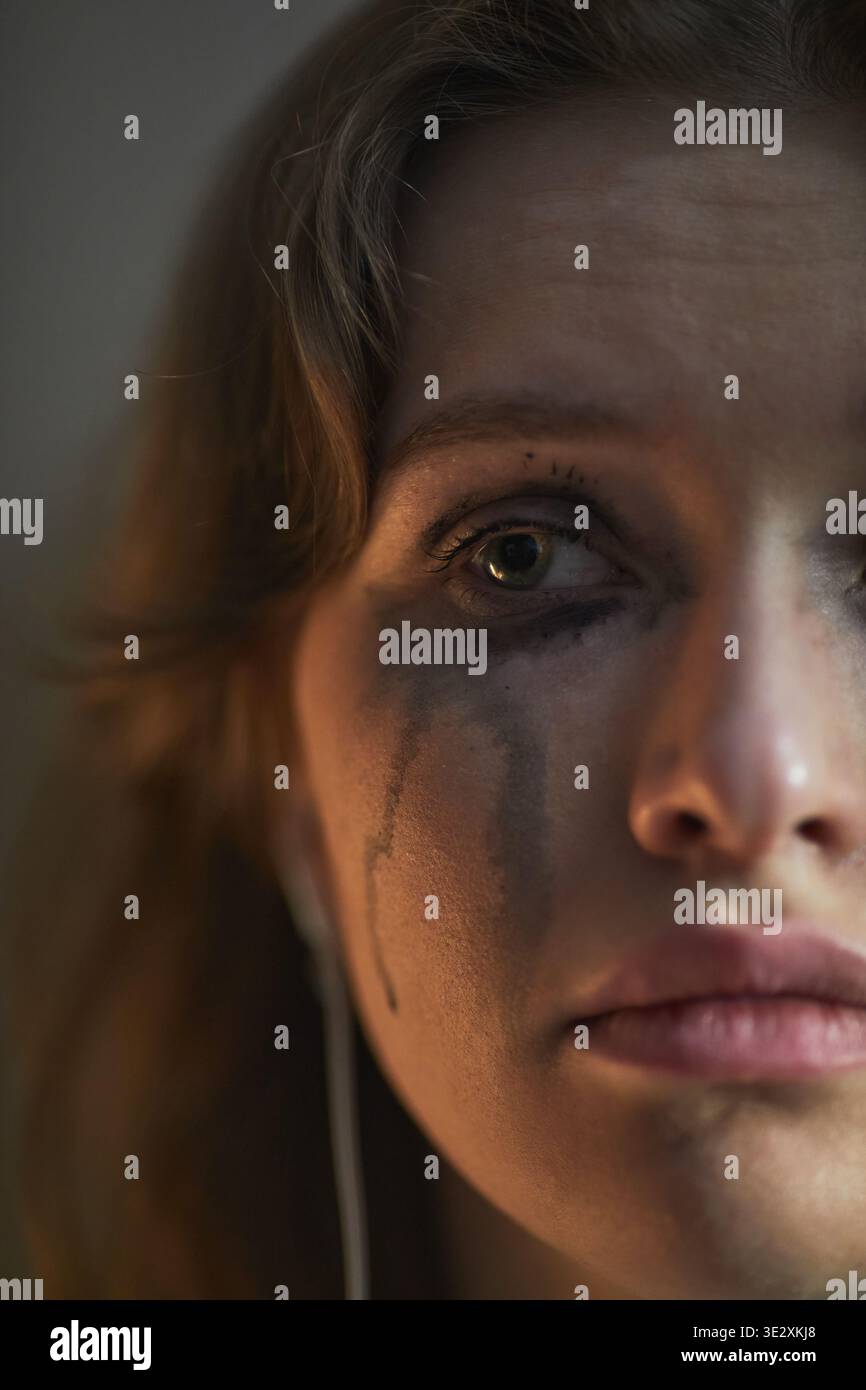 Vertical Portrait of young adult Caucasian woman looking into camera with tear-streaked face and smudged mascara, expressing intense sadness and vulnerability in close up composition Stock Photo