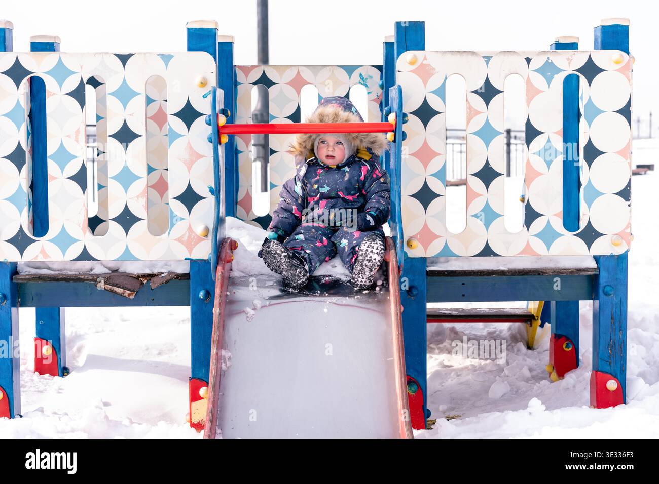A child in a colorful winter snowsuit slides down a snowy playground slide outdoors Stock Photo