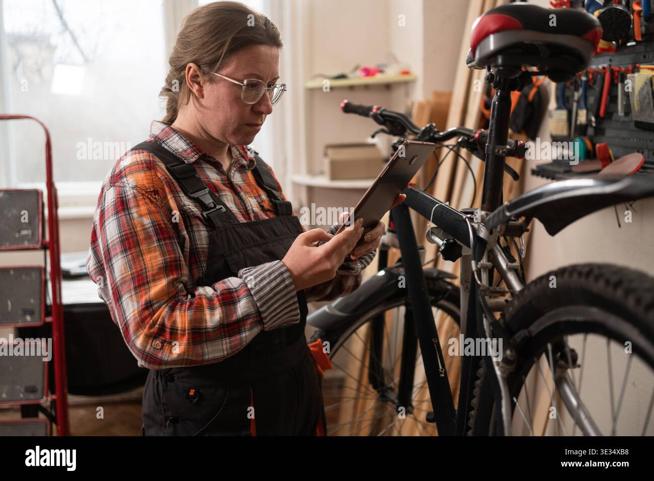 Woman bike mechanic using laptop for online diagnostics in workshop. Female technician searching for bicycle repair solution on internet. Concept of t Stock Photo