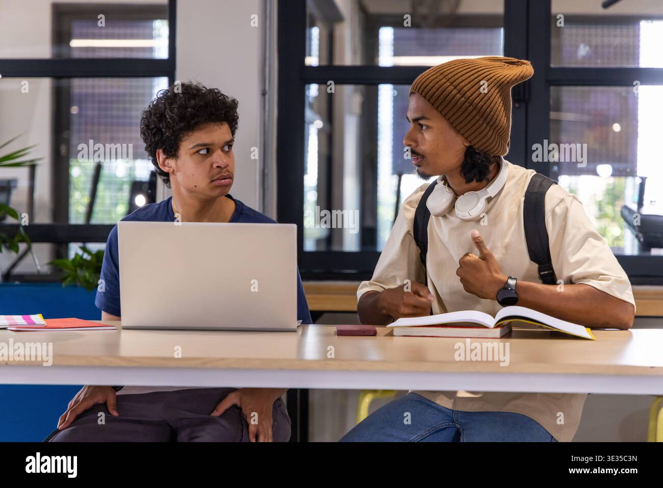 Diverse male students sitting at long wooden table studying with laptop, textbook and headphones Stock Photo