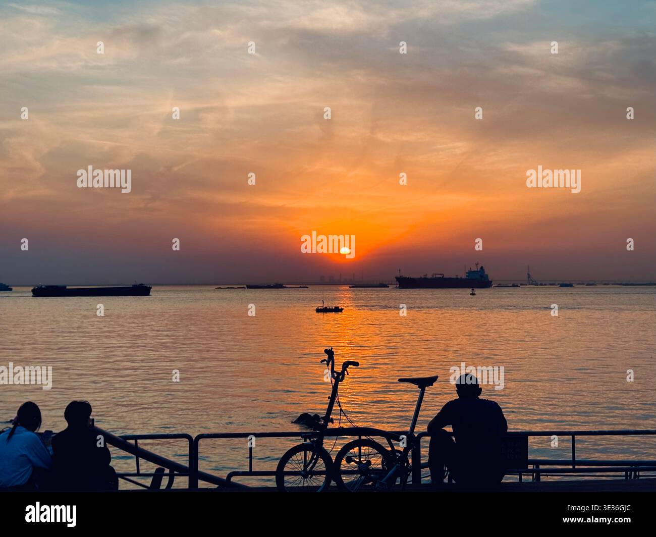 Golden sunset over the Yangtze River in Nantong, China. Silhouetted people and a bicycle on the riverfront as cargo ships pass in the distance. - Smartphone Captured Stock Image