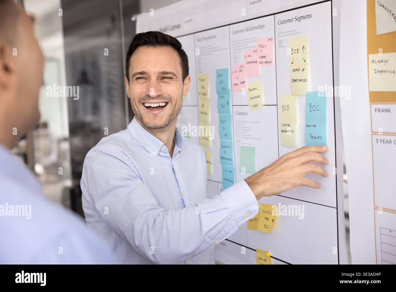 Laughing businessman showing male colleague sticky note on agile board Stock Photo