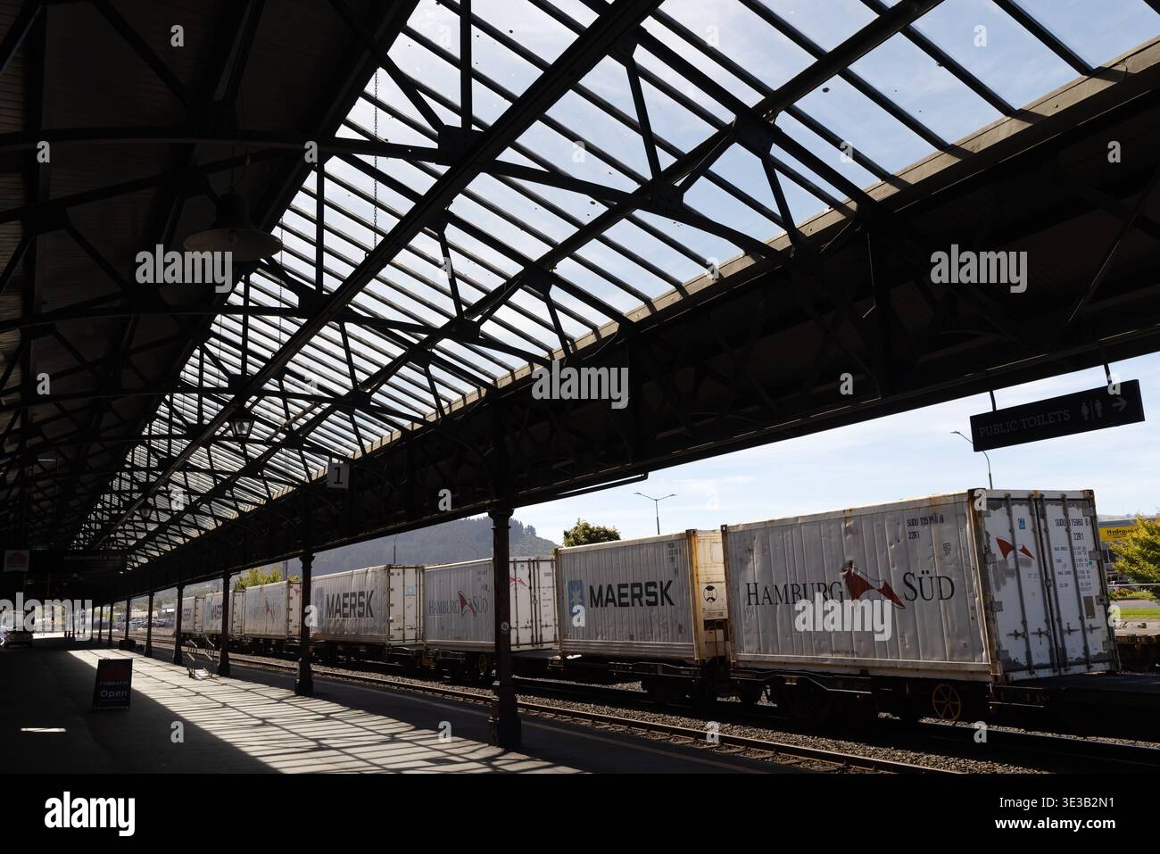 Freight train with shipping containers passes Dunedin Railway Station platform, Dunedin, Otago, New Zealand. Stock Photo