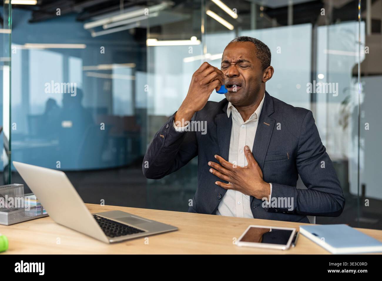 Mature businessman experiencing a respiratory attack, using an inhaler to alleviate breathing difficulties, placing a hand on his chest in an office setting Stock Photo