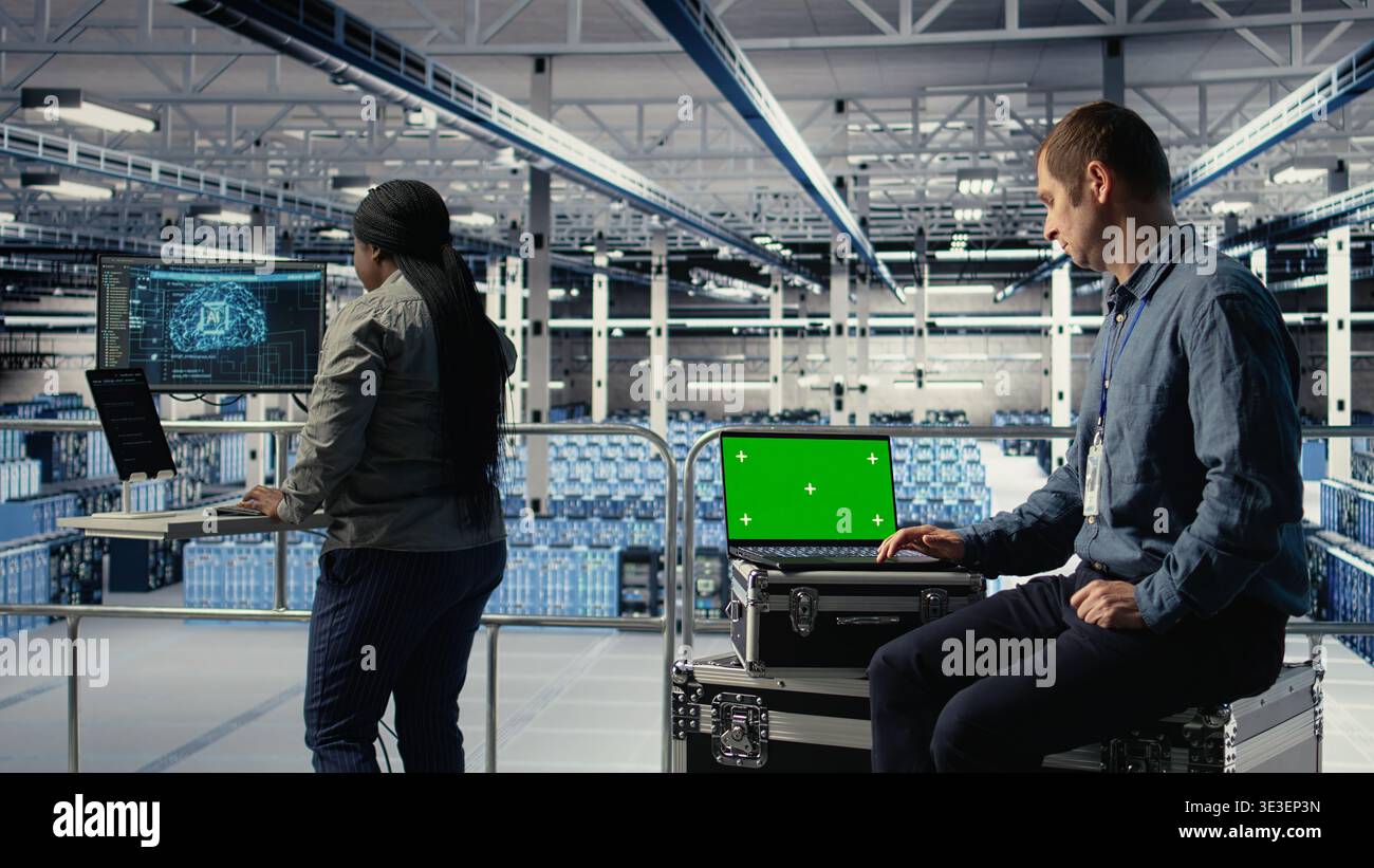 Data center IT experts investigate AI configurations using green screen laptop, making adjustments. Server hub engineers check deep learning errors on chroma key notebook, camera A Stock Photo