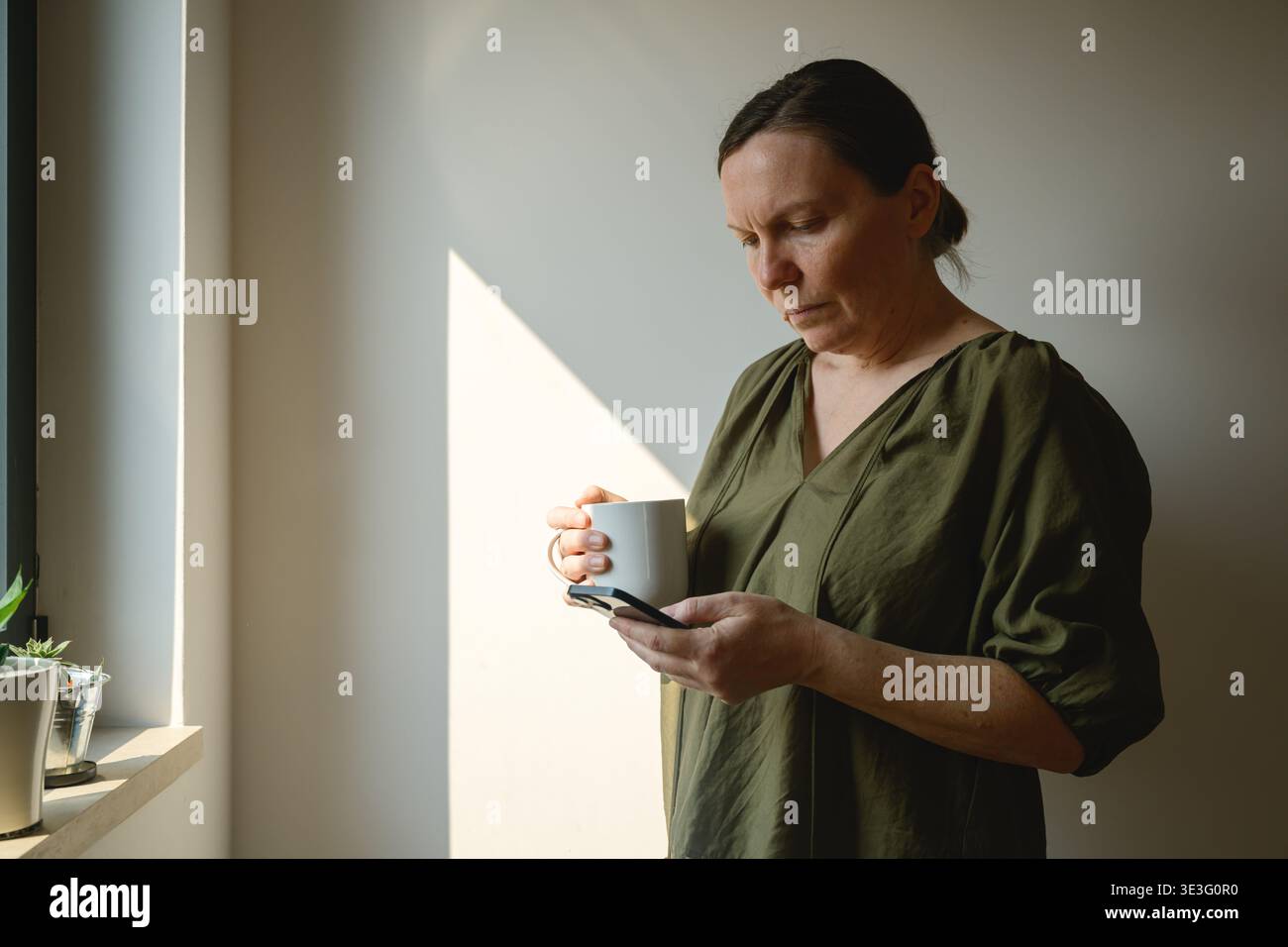 Mature woman using smartphone with coffee mug in hand near bright window, thoughtful expression in soft morning light at home, selective focus Stock Photo