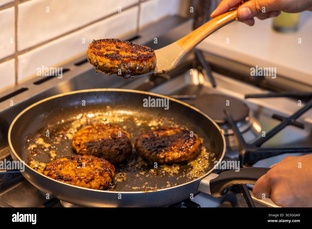 Home cook flipping seared burger patties in nonstick skillet on gas stove with wooden spatula Stock Photo