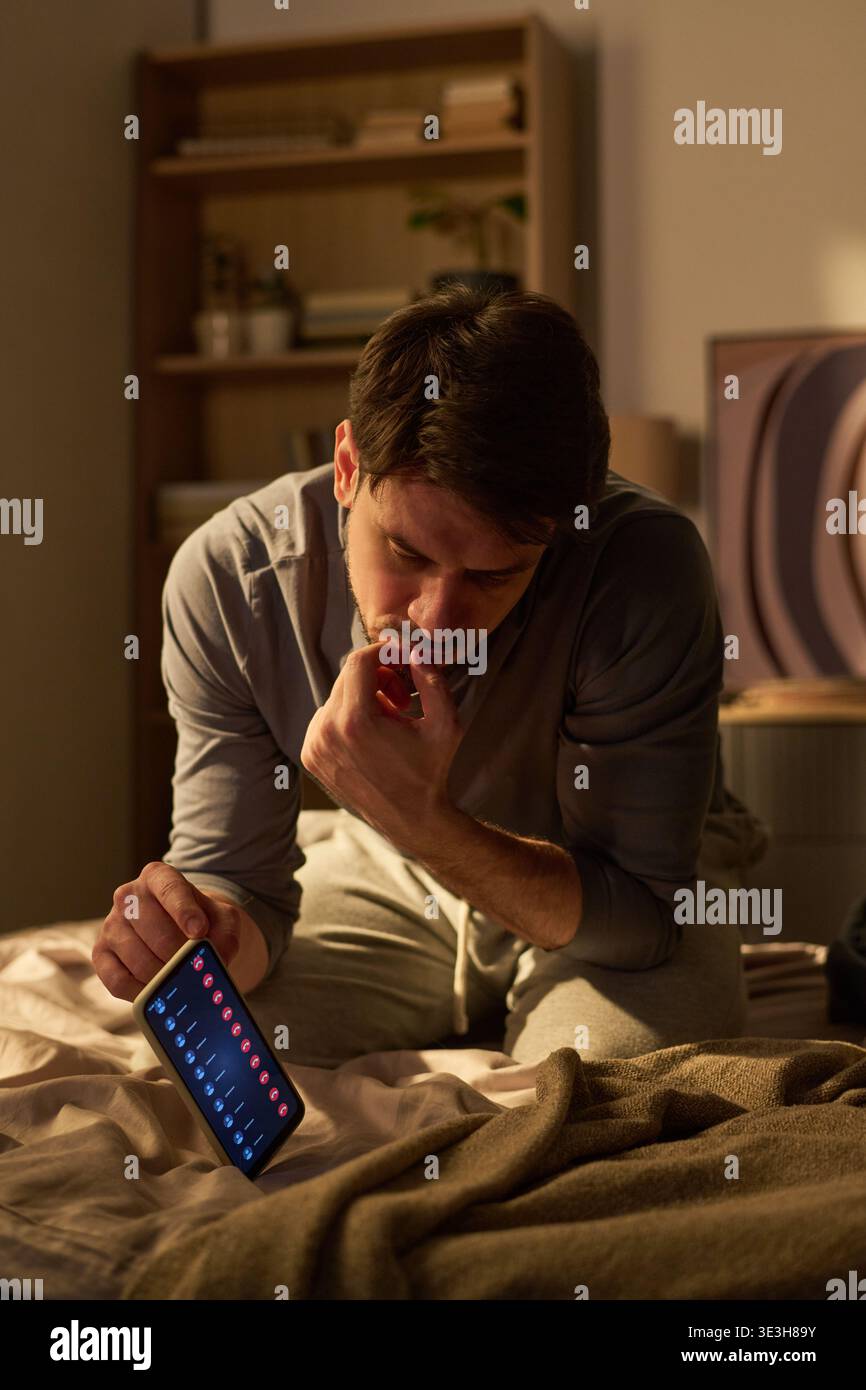 Young Man Sitting on Bed Checking Smartphone Notifications at Night Stock Photo