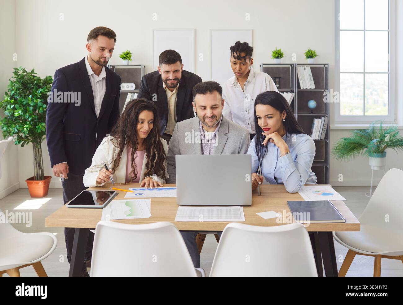 Diverse group, office professionals gathering around desk looking at laptop, document, discussing  Stock Photo