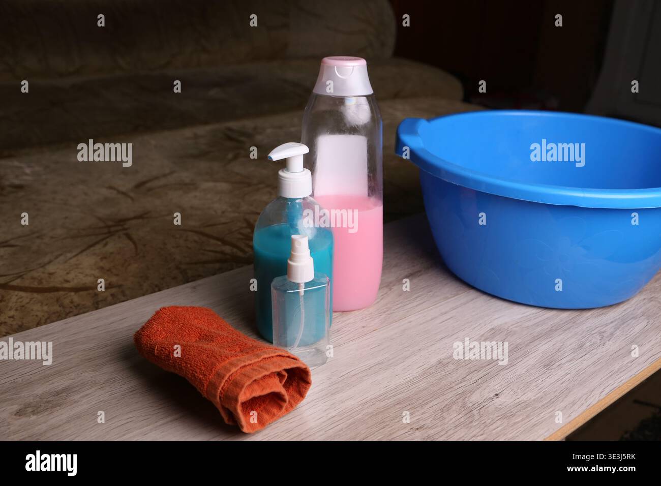 Cleaning supplies are placed on a table beside a blue bowl. There are bottles with different colored liquids and an orange towel ready for use in a cleaning task Stock Photo