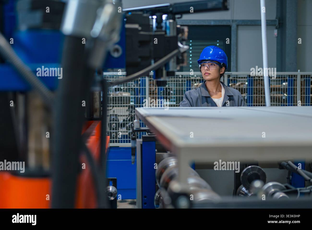 An Indian female engineer works in a factory wearing safety gear while operating machinery in a busy industrial environment. Stock Photo