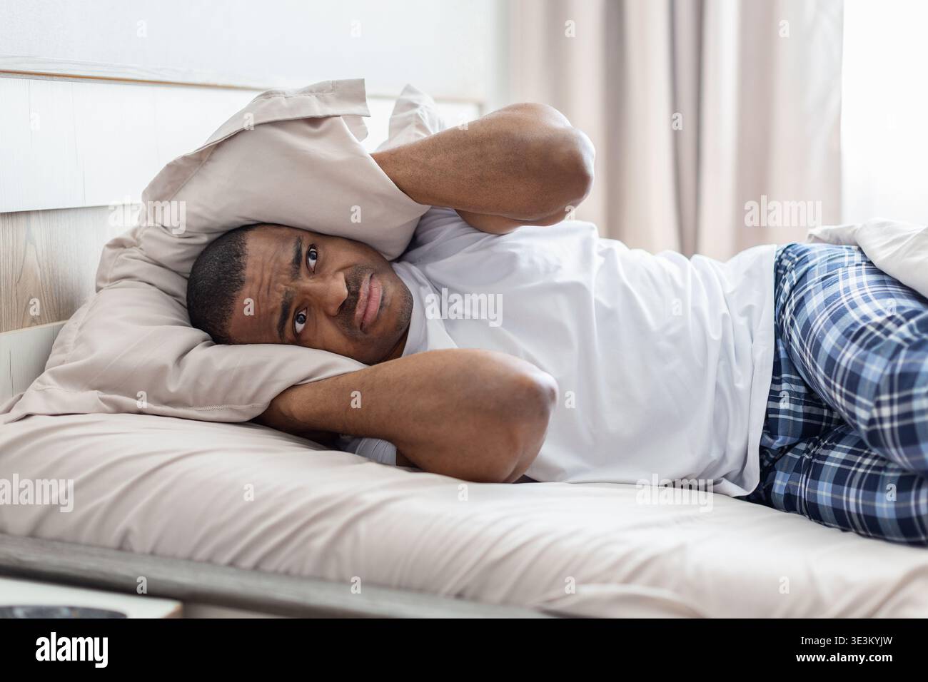 Young african american man lying in bed covering ears with pillow Stock Photo