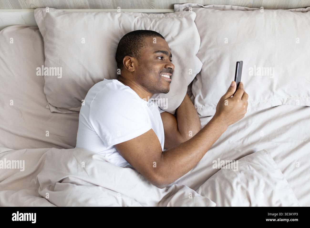 Black man lying in bed smiling while using smartphone, enjoying digital content Stock Photo