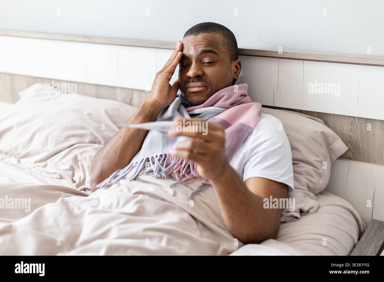 Young black man lying in bed checking temperature with thermometer Stock Photo