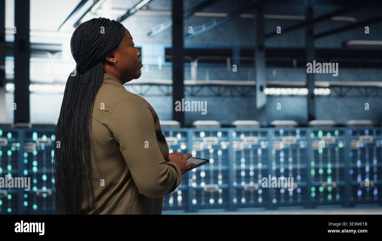 Data center programmer uses tablet to develop disaster recovery plans, minimizing downtime. IT professional walking in server farm, enhancing data protection protocols using device, camera B Stock Photo