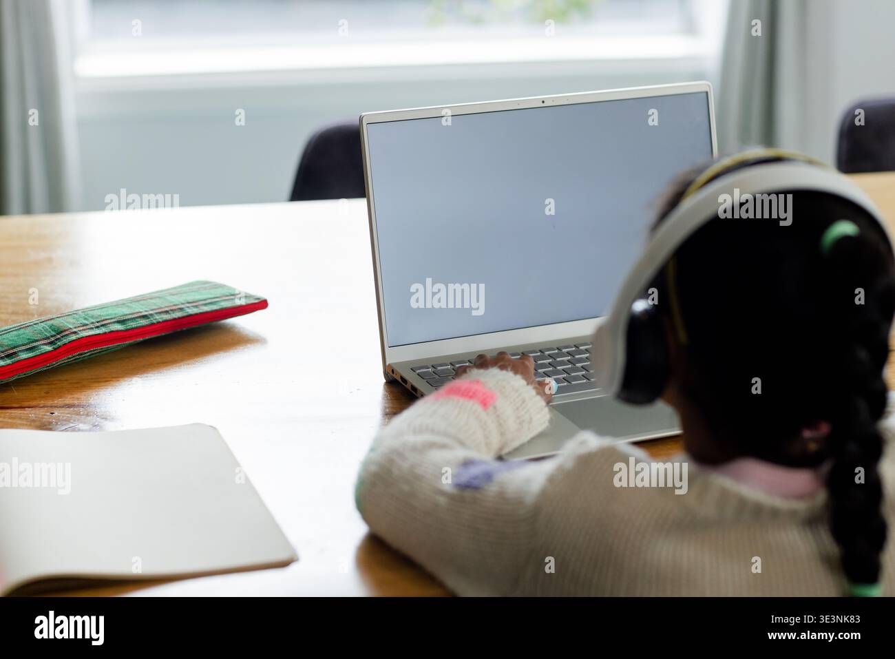 Child typing on laptop at home dining table near window, with notebook and patterned pencil case Stock Photo