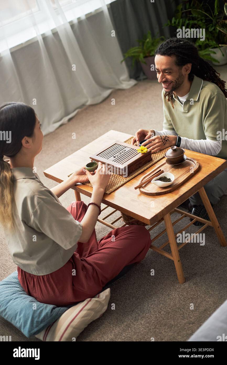 Asian woman and Black man sitting on floor playing traditional board game at low wooden table, both appearing as young adults, engaging in cultural activity with tea set visible Stock Photo