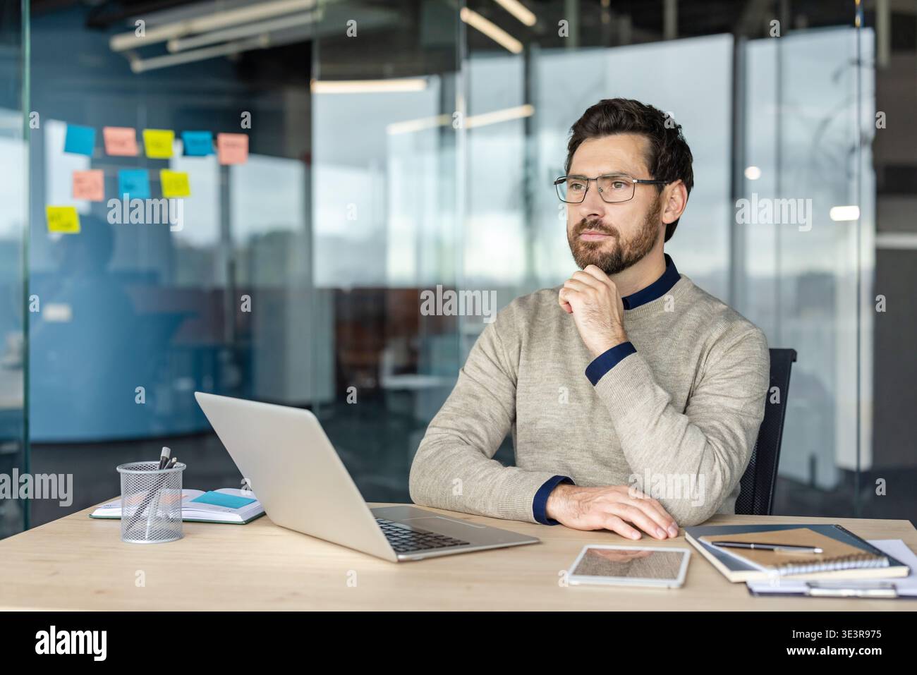 Man sitting at a modern office desk, resting his chin on his hand, engrossed in deep thought while planning future business strategies and solving problems Stock Photo