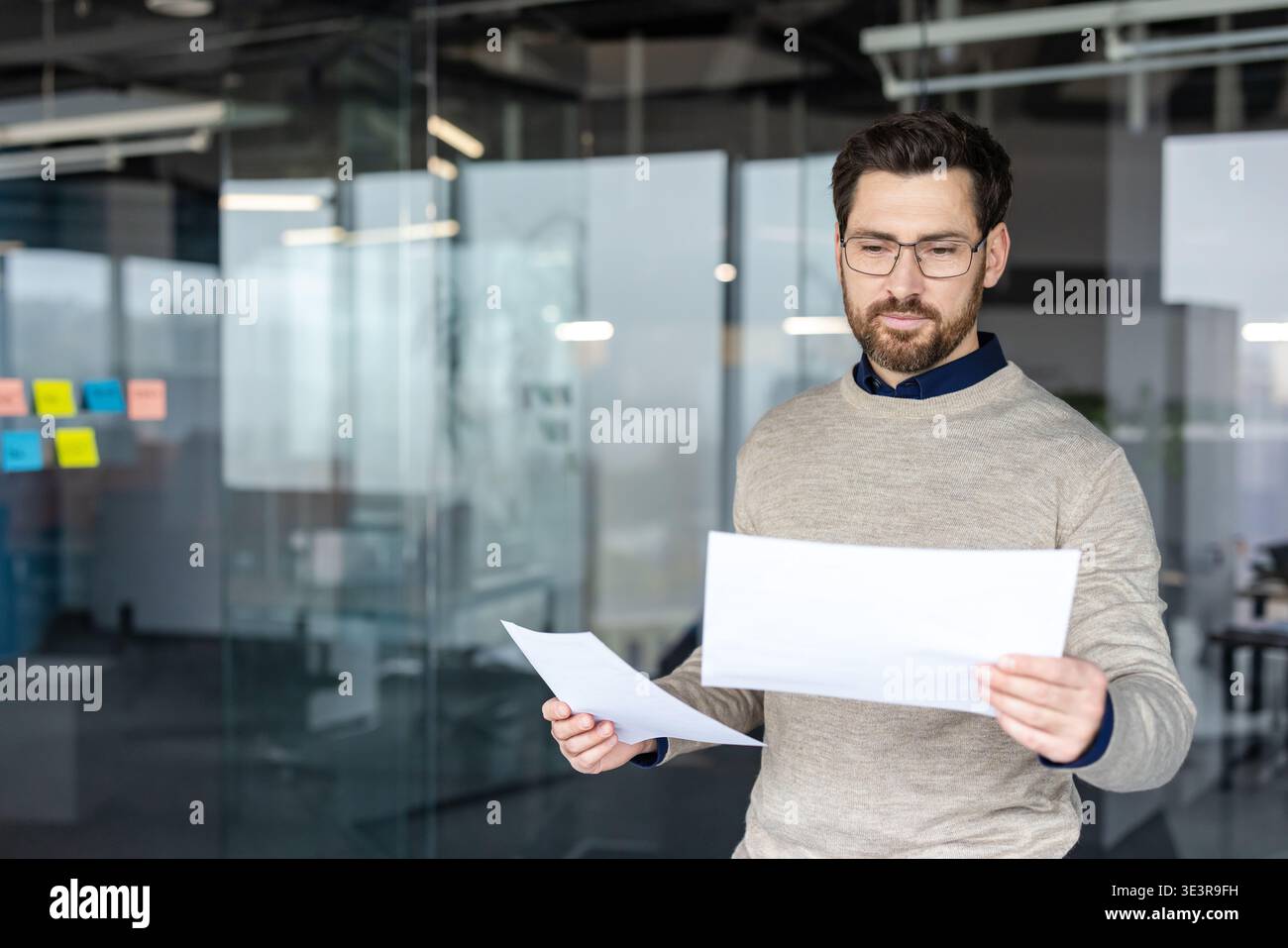 Young businessman standing in a contemporary glass office, reading and analyzing financial documents with a focused expression, representing data review and corporate management Stock Photo