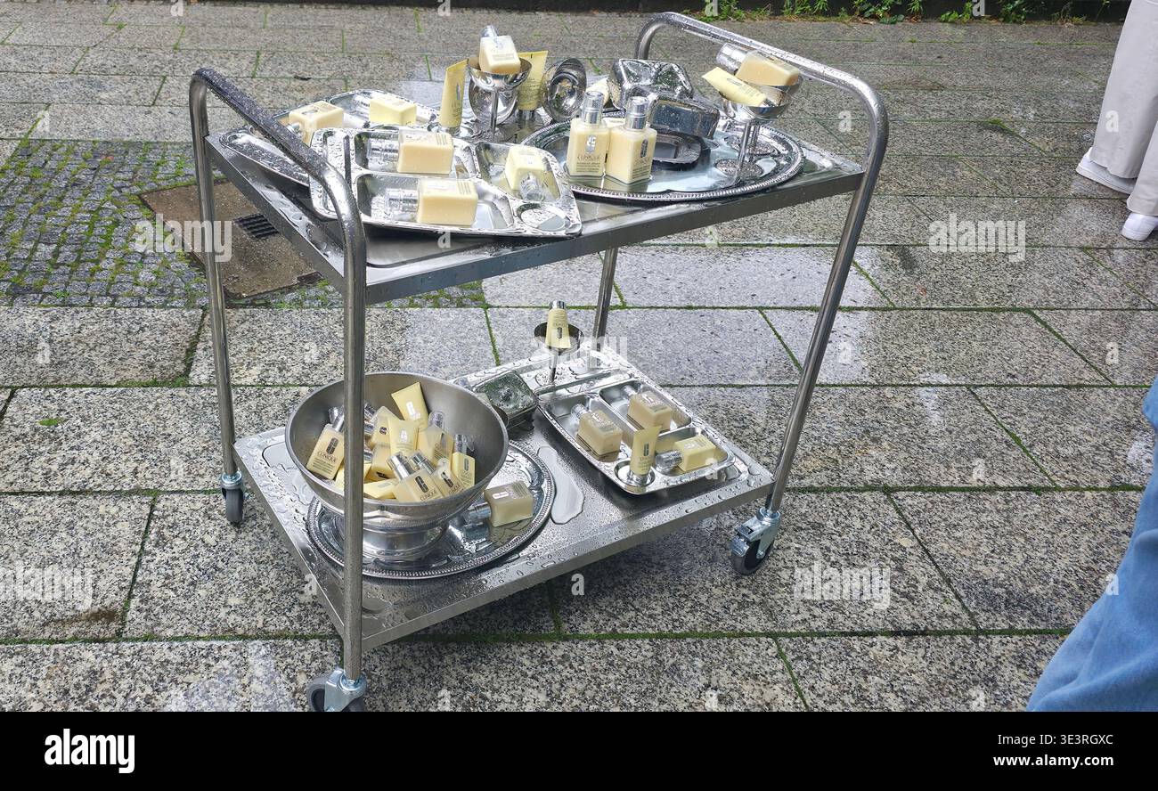 Berlin, Germany - July 17, 2025: . Metal serving cart displays various soap bars and trays on wet stone pavement - Smartphone Captured Stock Image