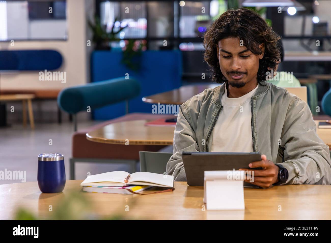 Indian man sitting at wooden table on right, using tablet on stand, notebook, notes, copy space Stock Photo