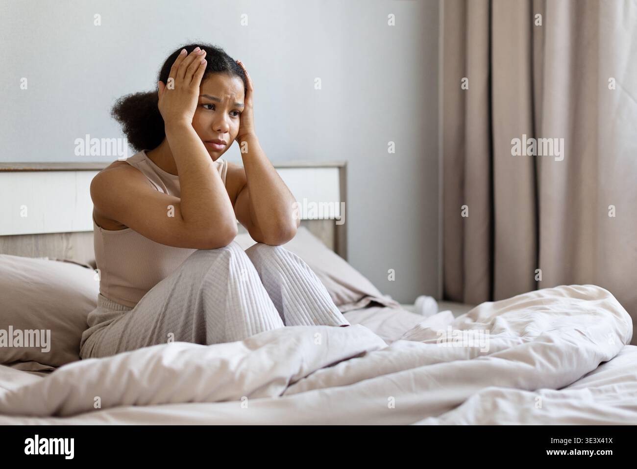 Stressed black woman sitting on bed holding her head with both hands Stock Photo