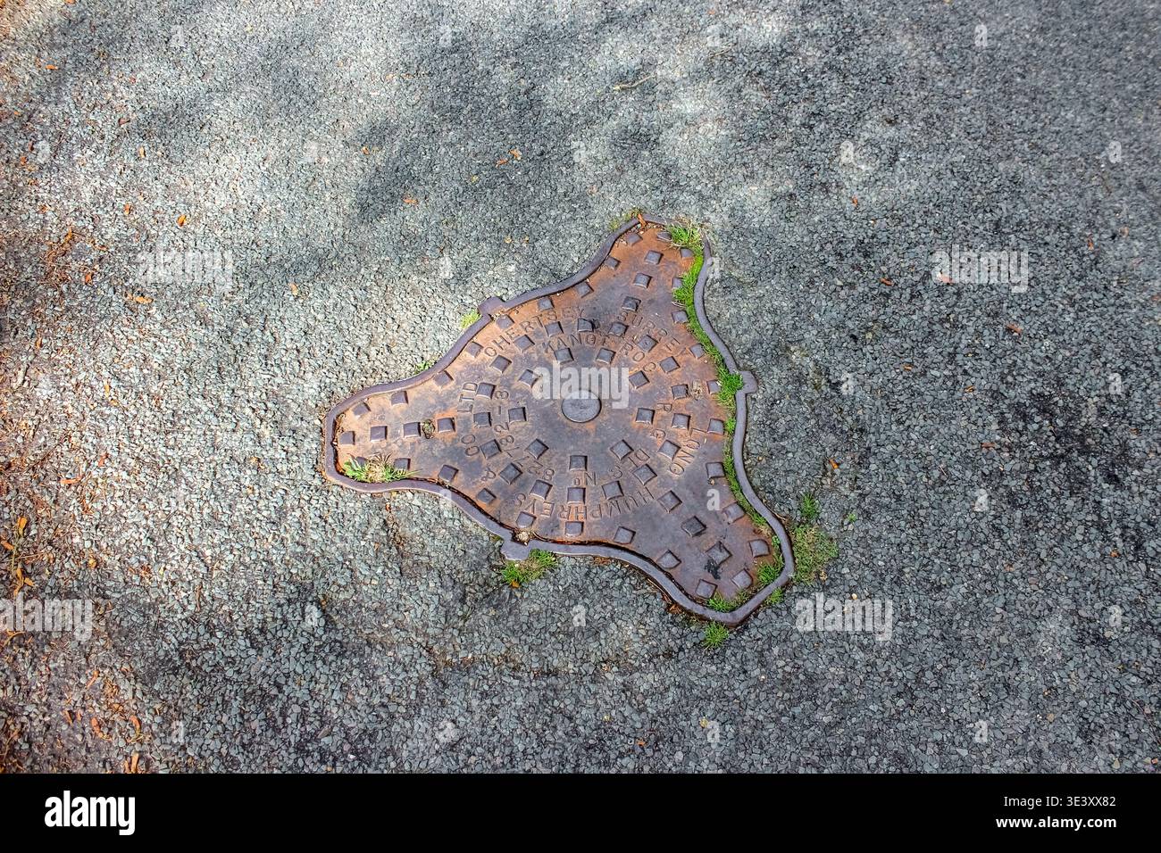 An aged, rusted triangular manhole cover with rounded edges is embedded in a weathered asphalt surface, featuring embossed text and square patterns. Stock Photo