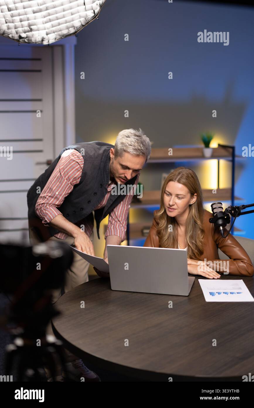 Podcasters reviewing printed analytics reports, collaborating using laptop in studio Stock Photo