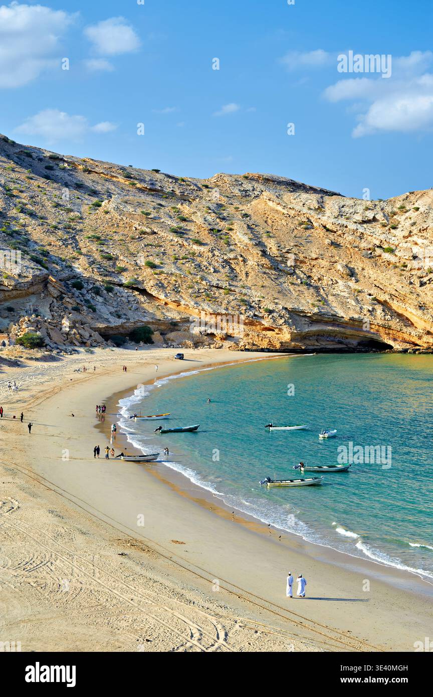 People walk along the sandy beach in Muscat, Oman. Fishing boats are present on the water, and hills rise in the background. Stock Photo