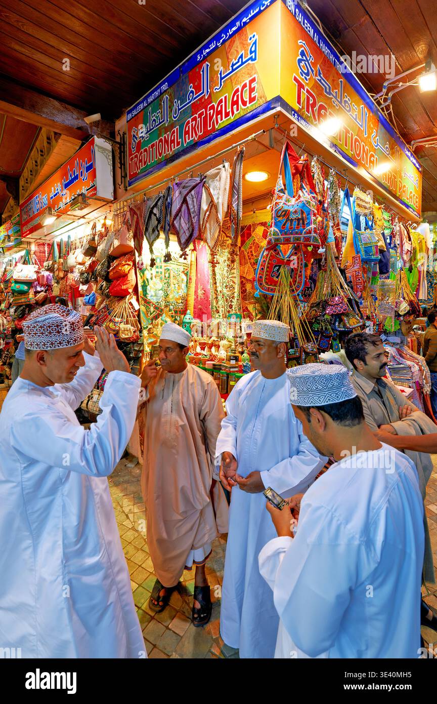 People gather outside shops in Mutrah Souq. Vendors display local crafts and goods while visitors share talks and explore the market. Stock Photo