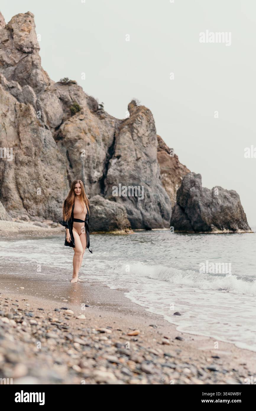 Woman beach cliffs walking on sandy shore with rocky mountains in background under overcast sky Stock Photo