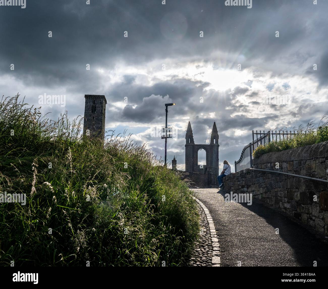 A lone person sits on a stone wall along the St Andrews esplanade, framed by the cathedral ruins and dramatic clouds as sunlight breaks through, creat Stock Photo