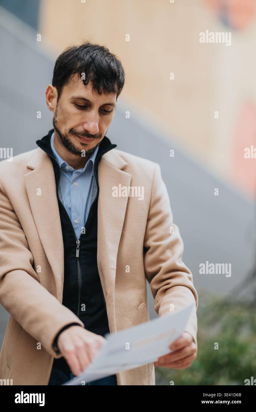 Businessman reading documents outdoors in a beige coat looking thoughtful during workday Stock Photo