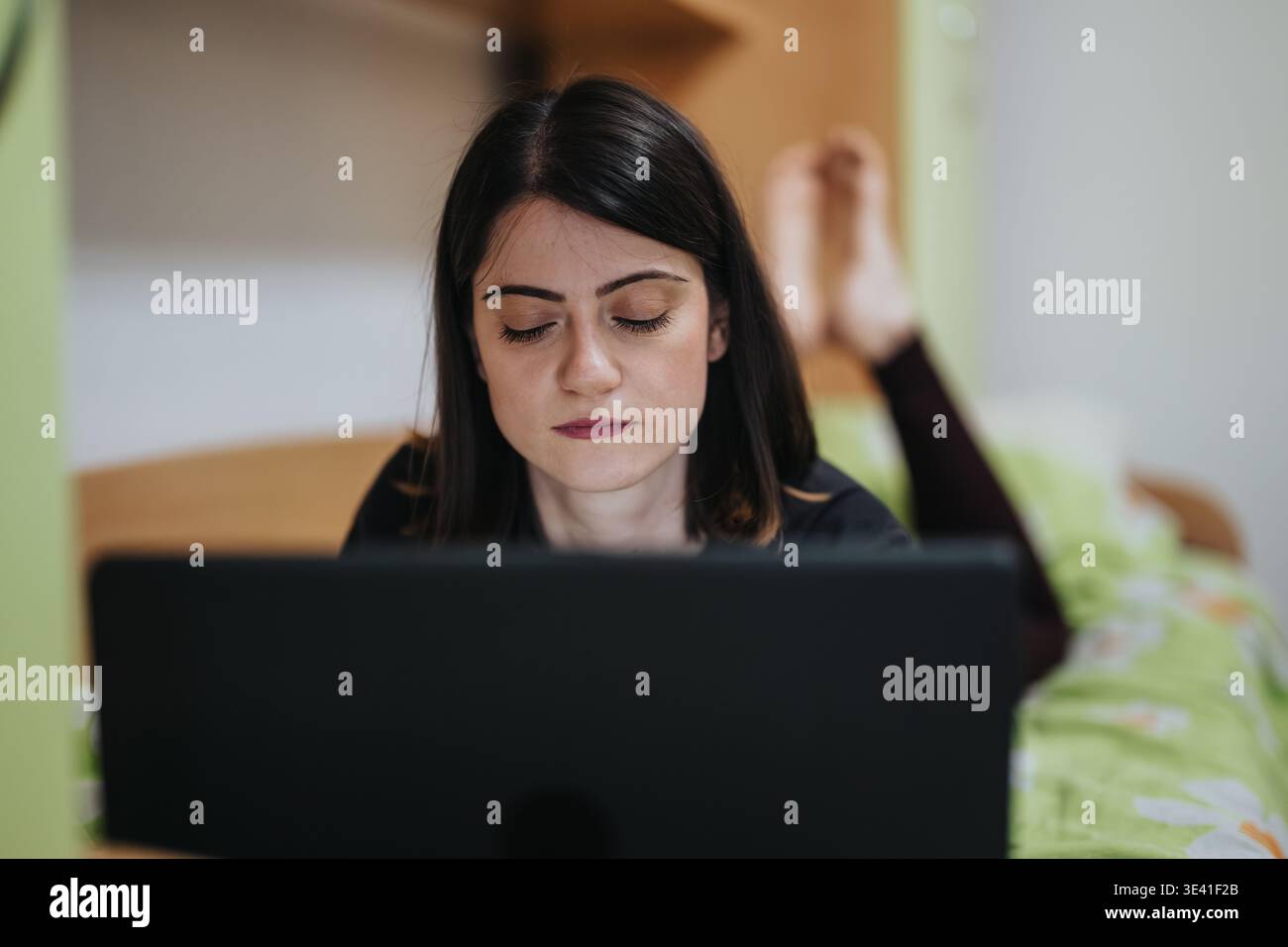 Young woman working on a laptop while lying on her bed at home, focused on the screen Stock Photo