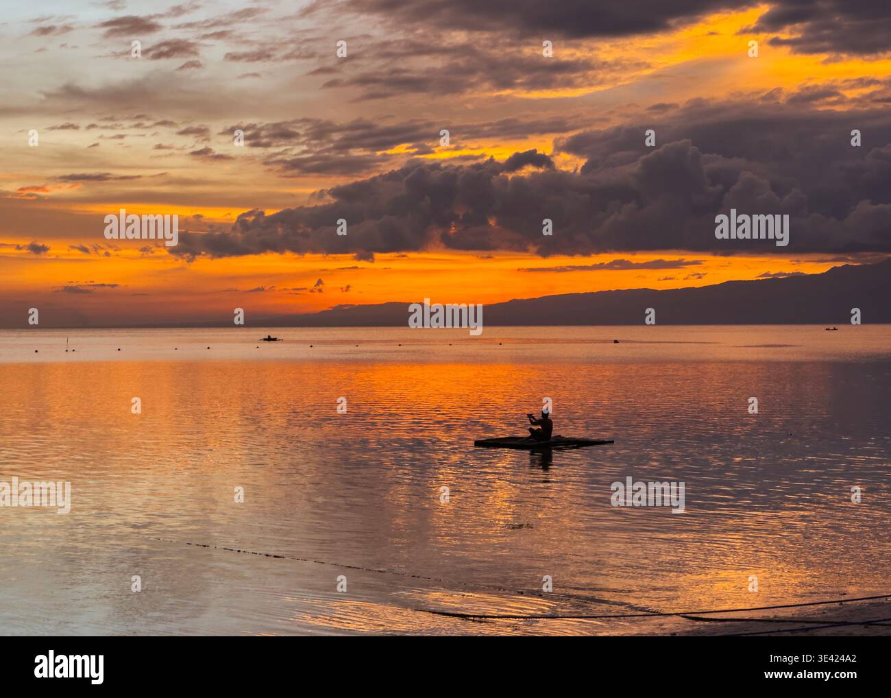 Silhouette of a man on a small raft at sunset on Little Paliton Beach, Siquijor, Philippines, with calm waters reflecting vivid orange skies. - Smartphone Captured Stock Image