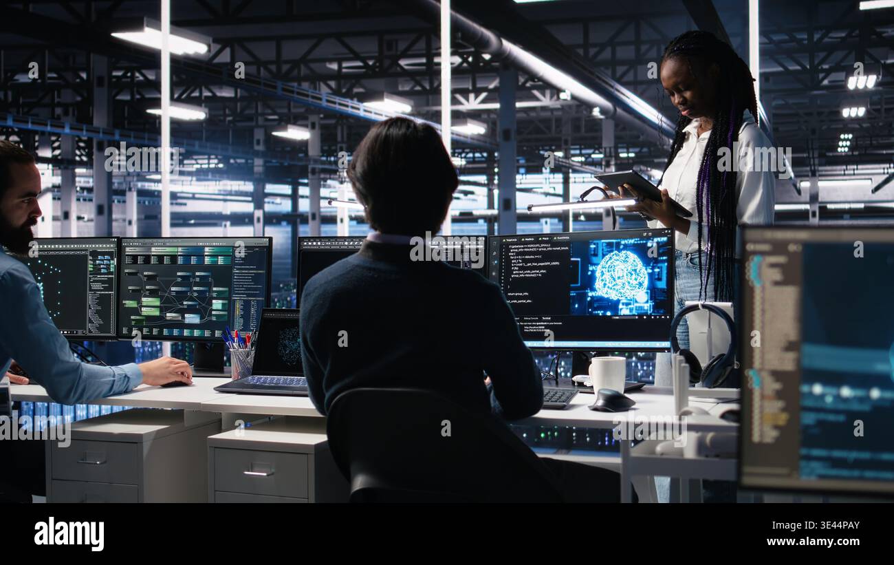 Technicians in high tech data center cooperate on neural network software development. Server hub IT workers talking, implementing deep learning technology using PC, camera A Stock Photo