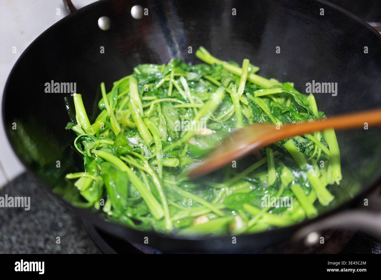 Stir-fried green vegetables being cooked in a black wok with wooden spoon, steaming and sizzling on Stock Photo