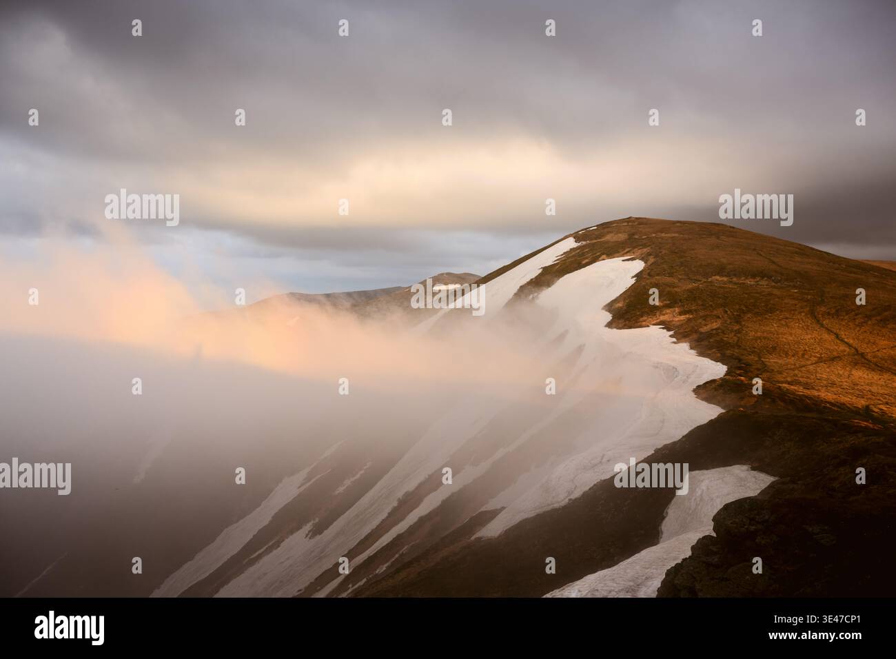 Mountain slope covered with snow streaks and rolling fog under cloudy sky. Misty atmosphere and dramatic terrain create cinematic wilderness mood Stock Photo