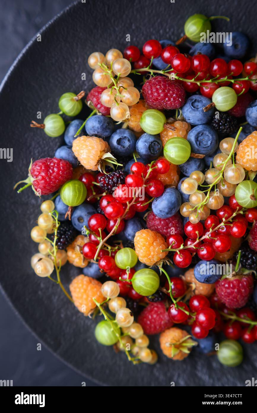Fresh assorted berries in black ceramic plate, minimal dark styling. Seasonal colorful fruit mix, healthy eating concept Stock Photo