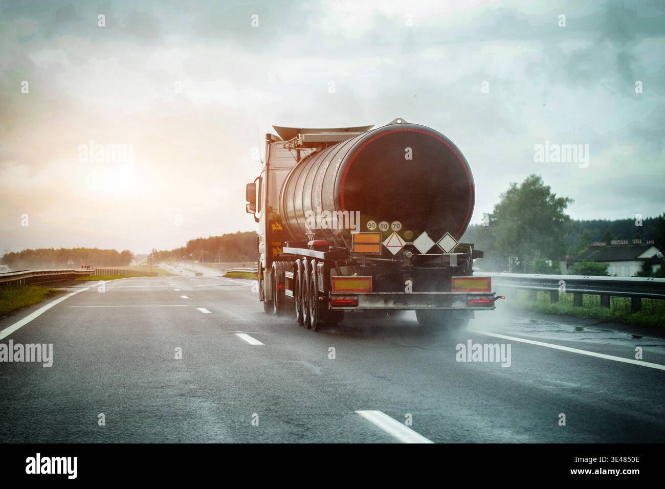 Rear view of tanker truck traveling on multilane highway during rainstorm, road surface wet with tire spray, overcast sky, hazardous material warnings Stock Photo