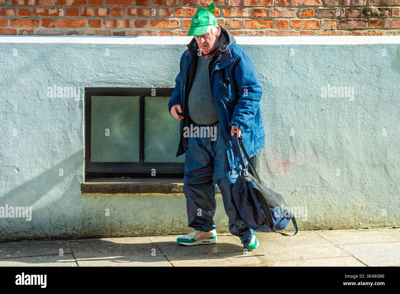Elderly local man in everyday attire wearing a shamrock cap walks through Dublin city centre on St Patrick’s Day subtly celebrating 17 March festival. Stock Photo